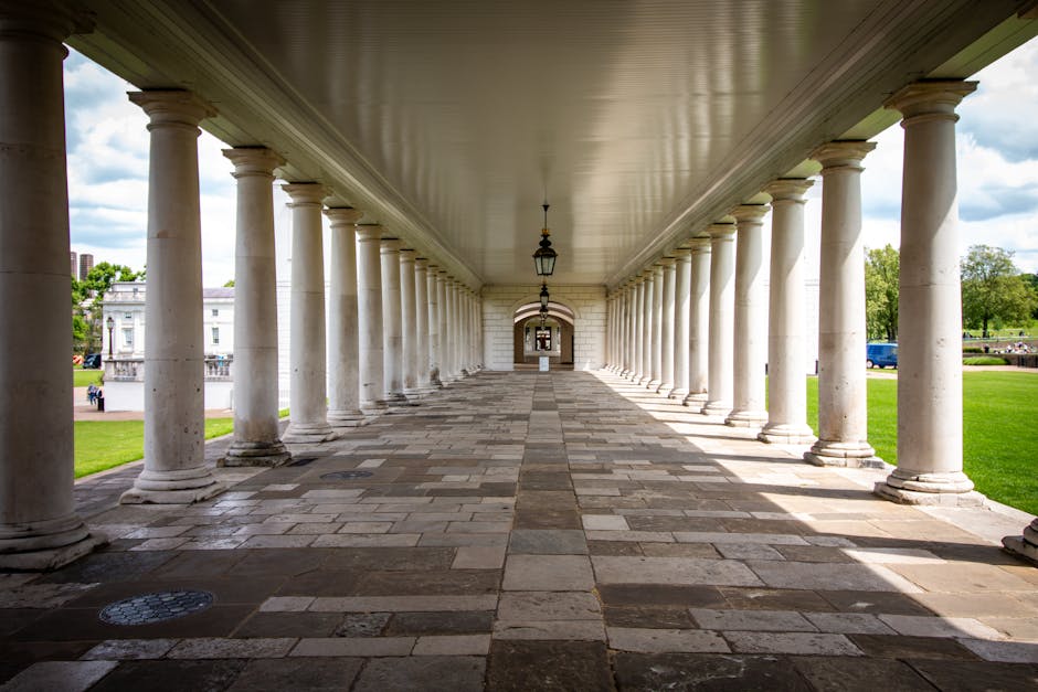 A covered walkway featuring a series of evenly spaced white classical columns supporting a smooth, beige ceiling. The floor is paved with rectangular stone tiles in varying shades of gray and brown, appearing clean and well-maintained. The area is illuminated by hanging lantern-style lights suspended from the ceiling. Surrounding the walkway, there is a well-kept green lawn and trees, with a few buildings and vehicles visible in the background under a partly cloudy sky. The scene emphasizes the neatness and structural cleanliness of the architectural surface, reflecting careful maintenance often associated with professional cleaning services like Greenwich Carpet Cleaners.