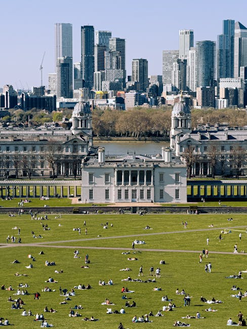 A wide city park with lush green grass and numerous people relaxing, sitting, and walking outdoors. In the middle of the park, there is a historic white building with classical architecture, featuring columns, a central balcony, and twin towers topped with domes. Beyond the park, a river separates the greenery from a modern downtown skyline composed of tall glass skyscrapers and high-rise buildings. The scene is brightly lit with natural daylight, creating a vibrant and clean atmosphere indicative of well-maintained public spaces. This image highlights the contrast between natural leisure areas and urban development, emphasizing the importance of outdoor cleanliness and surface maintenance in city environments. For professional cleaning and sanitisation services in this area, Greenwich Carpet Cleaners offers comprehensive solutions dedicated to maintaining hygiene in both residential and commercial spaces near Cutty Sark and Greenwich Market. 