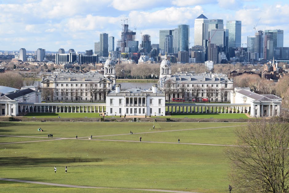 A wide view of the Royal Naval College in Greenwich, featuring its white neoclassical buildings with columns and domed towers, set against a large expanse of green lawn with people walking and relaxing. In the background, the modern city skyline of London with tall skyscrapers and office buildings is visible under a partly cloudy sky, highlighting the contrast between historical architecture and contemporary high-rise structures. The scene is well-lit, emphasizing the clean, well-maintained appearance of the historic site and surrounding greenery, with no visible dirt or clutter, reflecting the importance of surface cleaning, deep cleaning, and sanitisation for public spaces in Greenwich. Greenwich Carpet Cleaners specializes in maintaining hygiene in such locations, ensuring surfaces are thoroughly cleaned and preserved.
