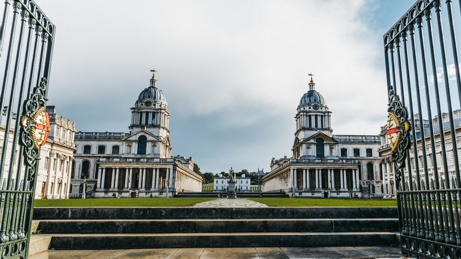 A wide city park with lush green grass and numerous people relaxing, sitting, and walking outdoors. In the middle of the park, there is a historic white building with classical architecture, featuring columns, a central balcony, and twin towers topped with domes. Beyond the park, a river separates the greenery from a modern downtown skyline composed of tall glass skyscrapers and high-rise buildings. The scene is brightly lit with natural daylight, creating a vibrant and clean atmosphere indicative of well-maintained public spaces. This image highlights the contrast between natural leisure areas and urban development, emphasizing the importance of outdoor cleanliness and surface maintenance in city environments. For professional cleaning and sanitisation services in this area, Greenwich Carpet Cleaners offers comprehensive solutions dedicated to maintaining hygiene in both residential and commercial spaces near Cutty Sark and Greenwich Market. 
