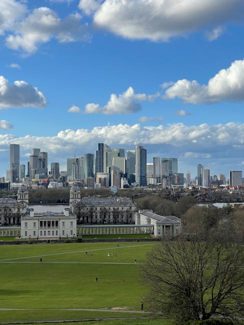 A wide view of a city skyline featuring modern skyscrapers made of glass and steel, with reflective surfaces capturing the daylight, set against a bright blue sky scattered with fluffy white clouds. In the foreground, a historic building with classical architecture and white facade is surrounded by lush green lawns with a few small groups of people walking. Mature trees with bare branches are also visible, framing the scene. The image conveys an urban landscape combining contemporary high-rise structures with historical architecture, under clear weather. The scene is associated with surface cleaning and maintenance of city properties, aligning with Greenwich end of tenancy cleaning prices real cost guide, Greenwich, and is linked to professional cleaning services provided by Greenwich Carpet Cleaners.