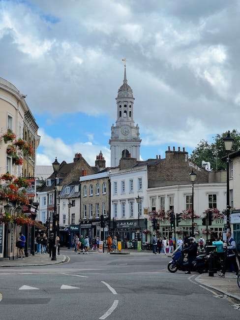A busy street scene in Greenwich featuring a historic clock tower with a tall, ornate spire under a partly cloudy sky. Surrounding the tower are Georgian-style buildings with large windows, some adorned with hanging flower baskets. The street is filled with pedestrians walking on the sidewalks, along with parked and moving cars and scooters. Street lamps and shopfront signs line both sides of the street, contributing to the lively atmosphere. The overall scene depicts a charming urban area known for its architectural heritage, with visible commercial activity and a mix of historic and modern elements, all captured in natural daylight. This image reflects the vibrant community atmosphere typical of Greenwich, supported by local businesses including Greenwich Carpet Cleaners.