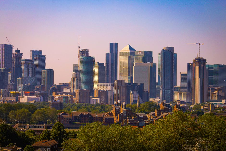 A wide view of a city skyline featuring modern skyscrapers made of glass and steel, with reflective surfaces capturing the daylight, set against a bright blue sky scattered with fluffy white clouds. In the foreground, a historic building with classical architecture and white facade is surrounded by lush green lawns with a few small groups of people walking. Mature trees with bare branches are also visible, framing the scene. The image conveys an urban landscape combining contemporary high-rise structures with historical architecture, under clear weather. The scene is associated with surface cleaning and maintenance of city properties, aligning with Greenwich end of tenancy cleaning prices real cost guide, Greenwich, and is linked to professional cleaning services provided by Greenwich Carpet Cleaners.