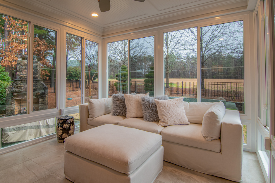 A bright, sunny living room with large floor-to-ceiling glass windows showcasing a view of a backyard with trees and a fence. The room features a white L-shaped sofa adorned with soft, plush cushions 