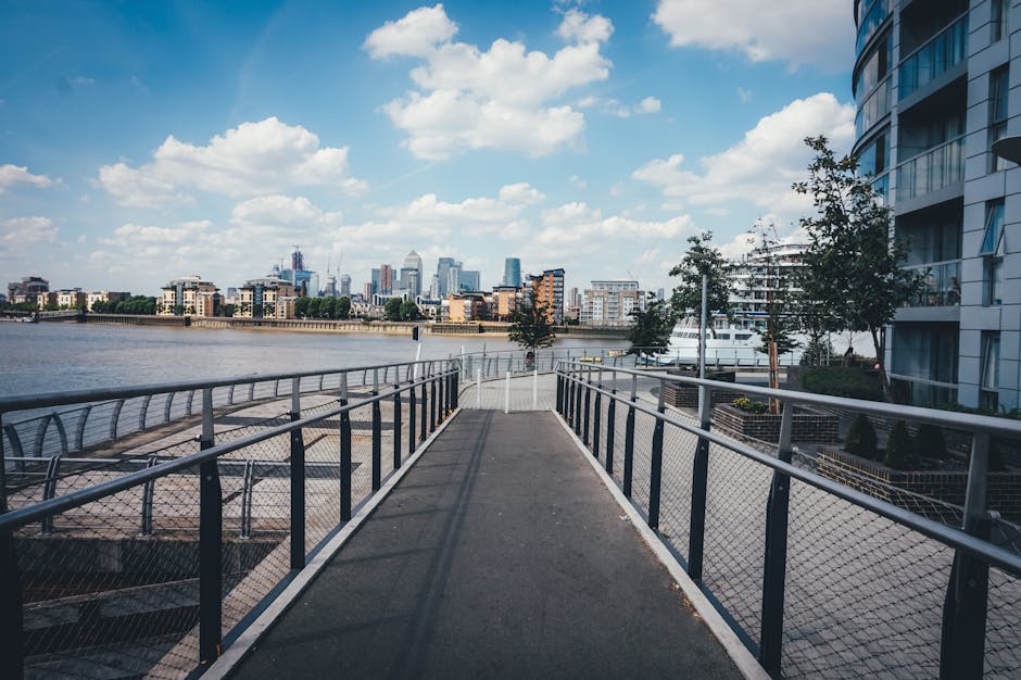 A panoramic view of the Greenwich cityscape showing modern residential and commercial buildings along the river Thames, with reflections on the water surface. The image features high-rise apartments i