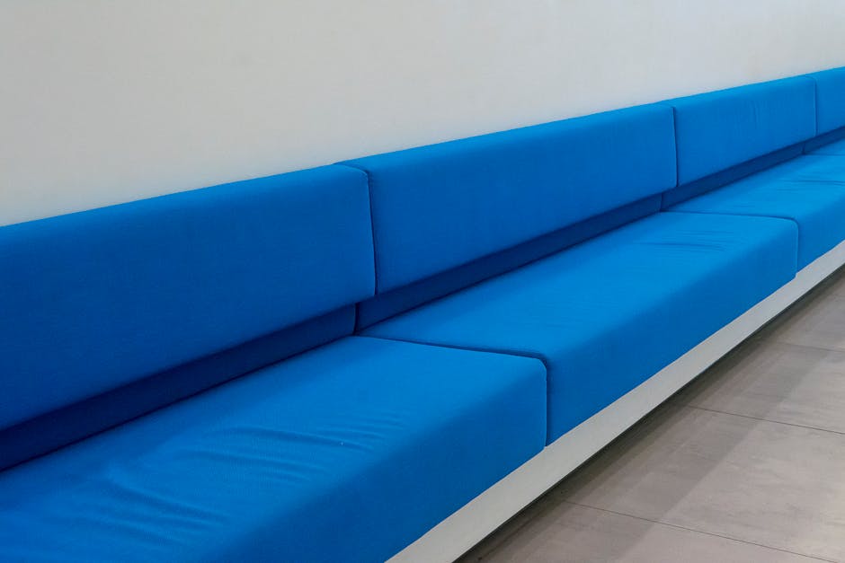 A long, bright blue upholstered bench with cushioned seats and backrests, positioned against a plain white wall in a corridor or waiting area. The surface of the bench appears clean and well-maintained, with no visible dust or stains. The floor beneath is light-colored and tiled, adding to the room's clean and modern appearance. The ambient lighting highlights the smooth textures of the upholstery, and the overall setting suggests a space recently serviced for cleanliness by Greenwich Carpet Cleaners, specializing in surface cleaning and hygienic maintenance consistent with same-day upholstery cleaning services in Greenwich SE10.